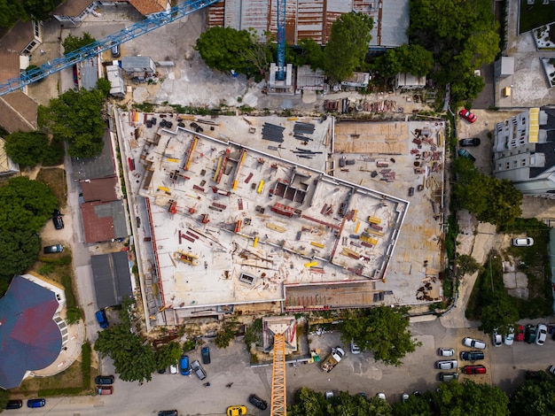 An aerial shot of a construction site in Medellín, Colombia. The site is bustling with activity as workers operate heavy machinery, laying the foundation for a new residential building. The surrounding cityscape includes lush green hills and other ongoing development projects.